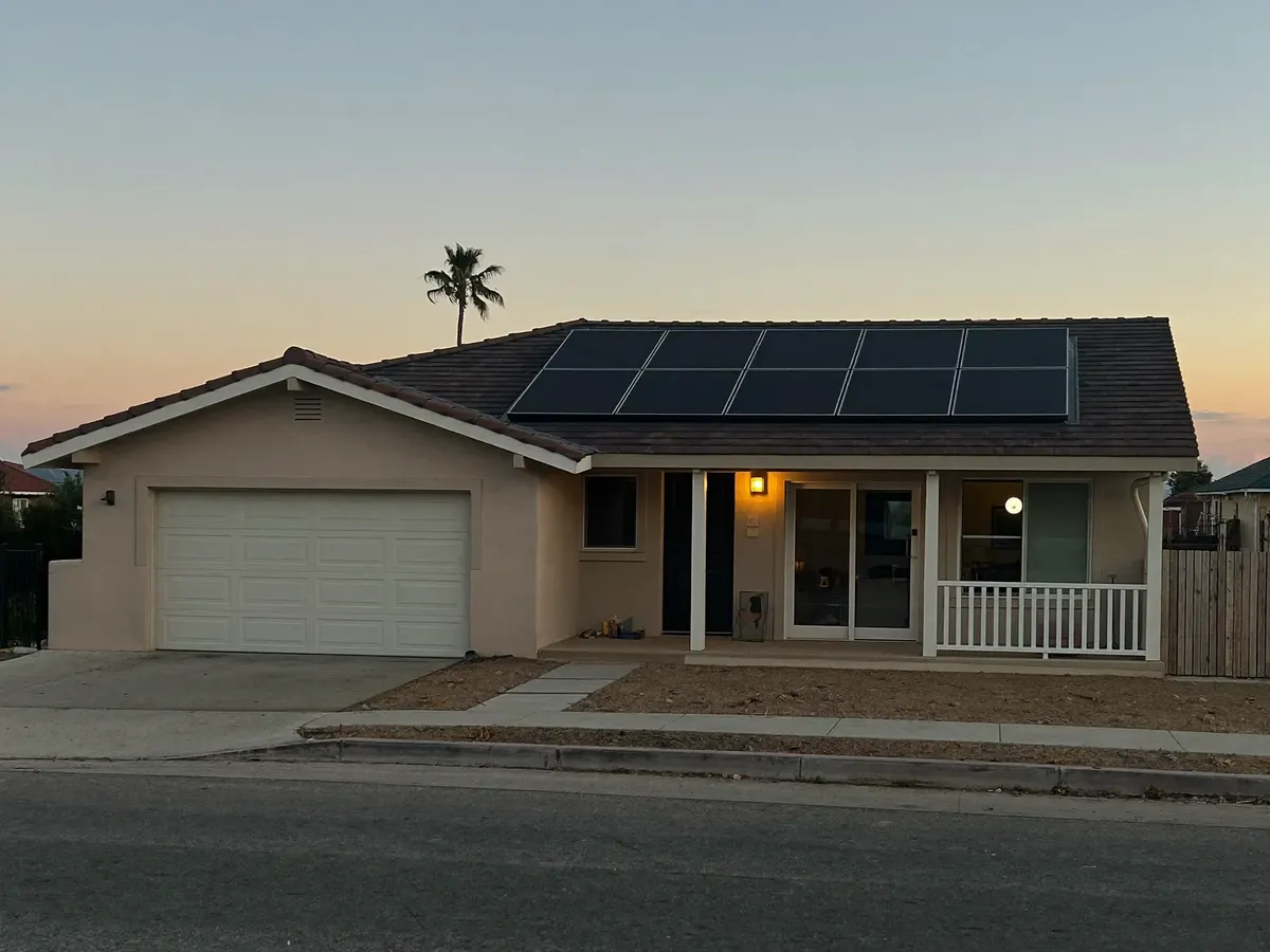 A single-story stucco home with solar panels on its tile roof at dusk, porch light on.