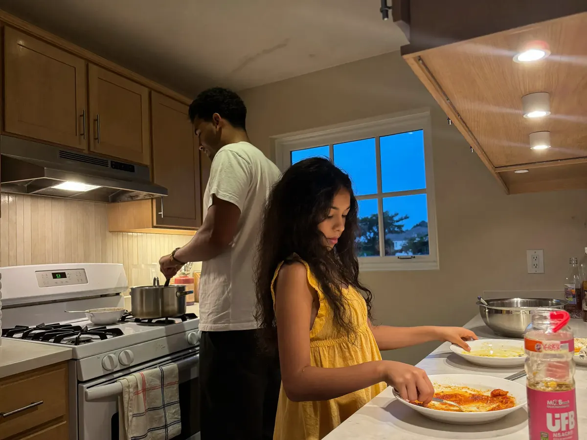 A mother cooks dinner while her teenage son sets plates on the counter in a California kitchen in the early evening.
