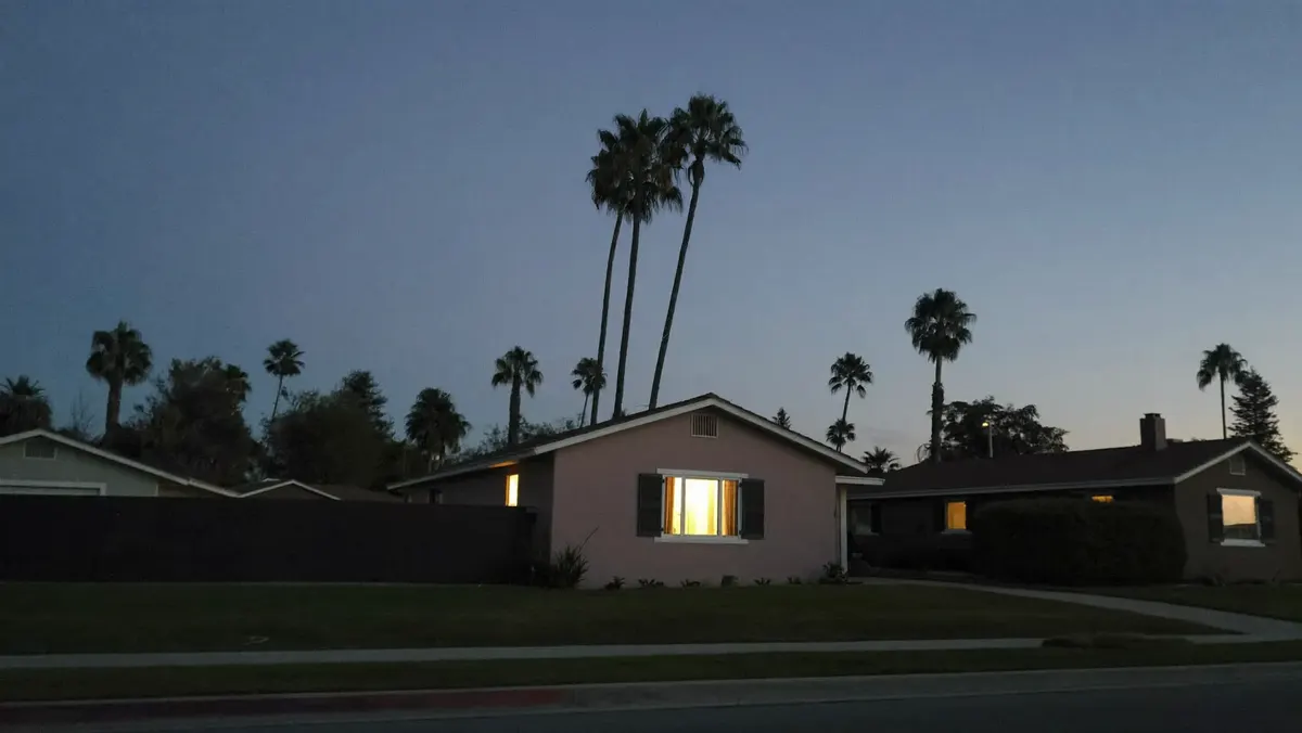 A quiet California street at dusk with the houses dark except one with its windows lit.